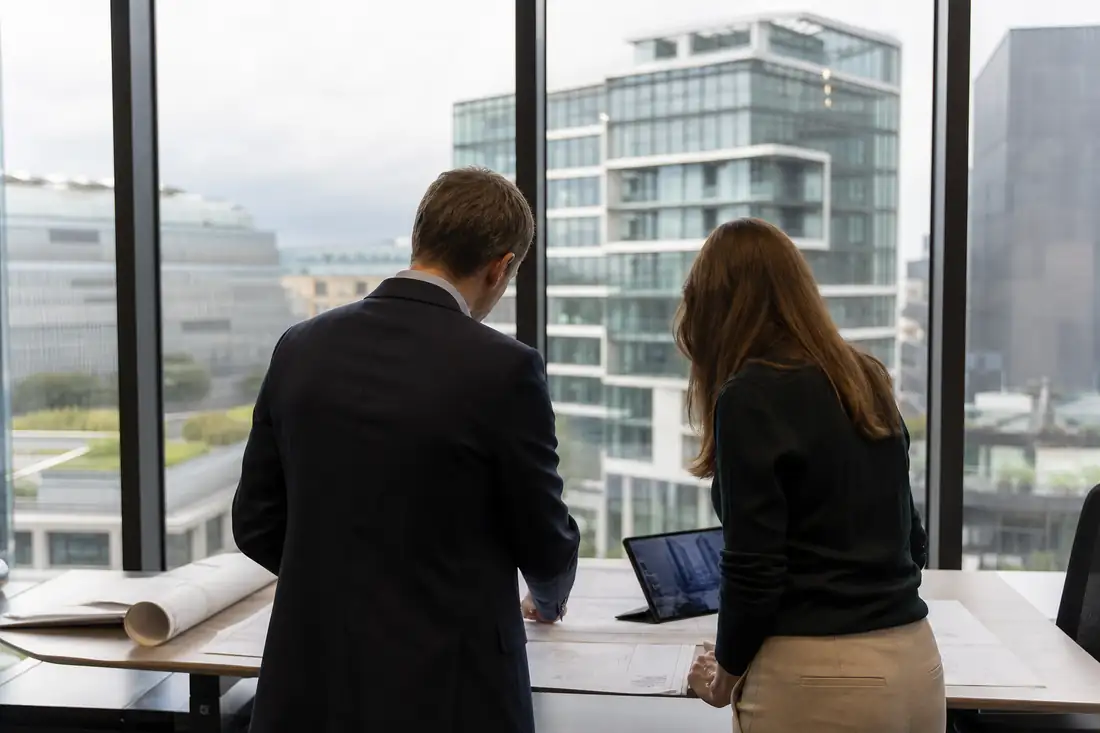Deux professionnels de l’immobilier examinent des plans devant des immeubles de bureaux contemporains.