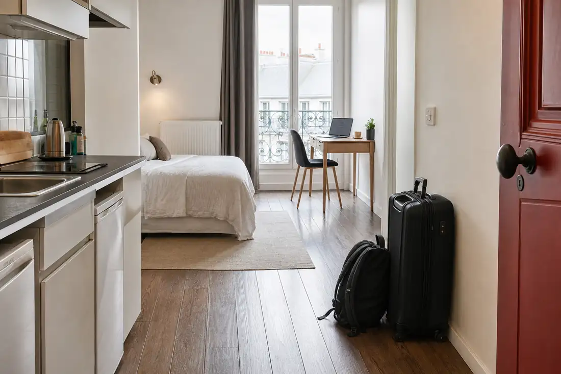 Studio meublé lumineux avec une valise et un sac à dos près de l’entrée, lit fait et bureau devant la fenêtre