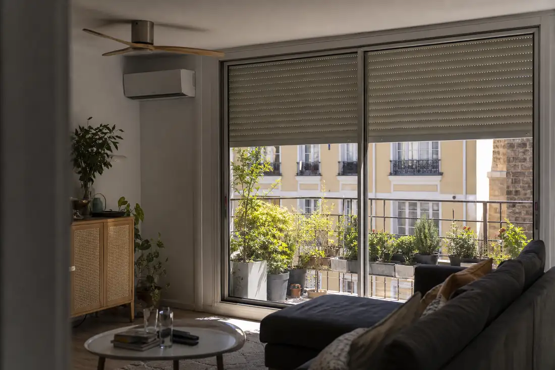 Salon d’appartement protégé du soleil par des volets extérieurs, avec ventilateur de plafond et climatisation murale.