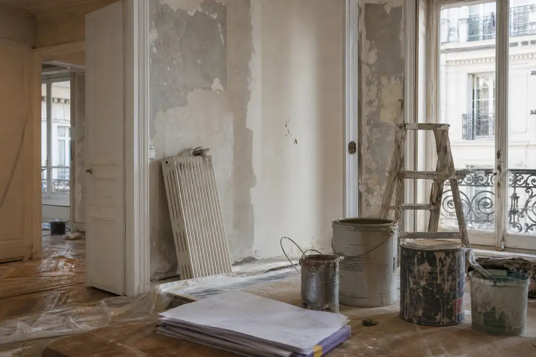 Appartement ancien en cours de rénovation, murs décapés, radiateur déposé, escabeau et pots de peinture au sol.
