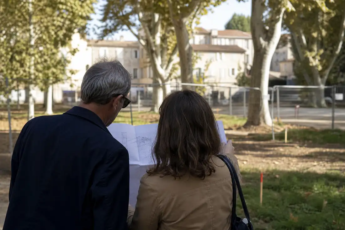 Parc arboré à Narbonne bordé par une emprise de chantier et des habitants consultant un plan d’urbanisme.