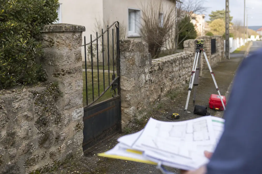 Mur ancien et matériel de géomètre matérialisant la limite incertaine entre deux jardins résidentiels.