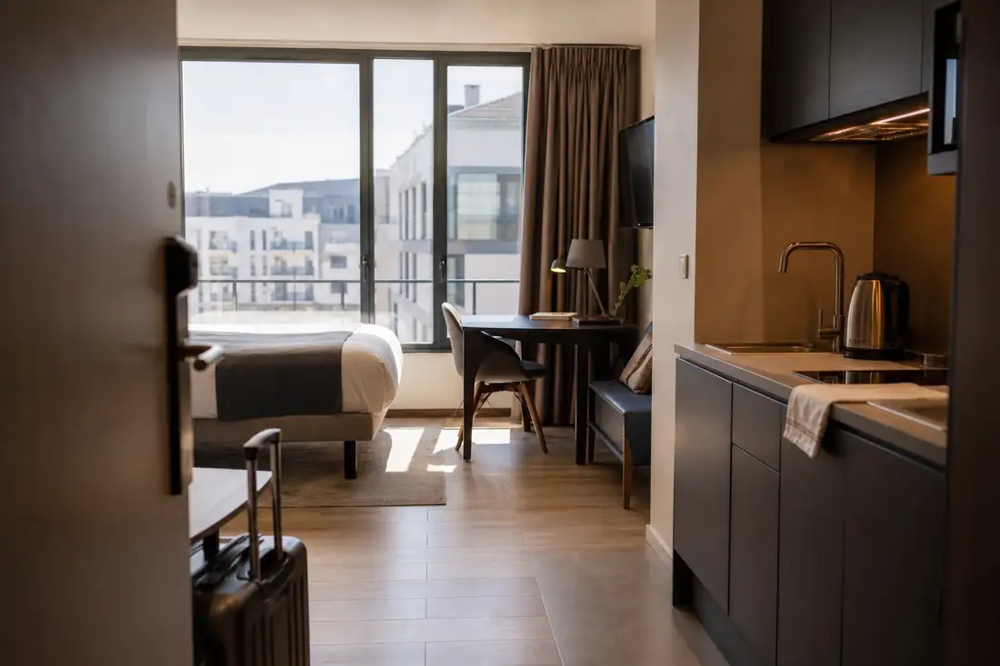 Studio meublé d’un aparthotel francilien avec kitchenette, bureau et lumière matinale sur un quartier urbain.