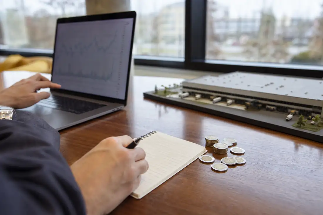 Bureau d’investisseur avec ordinateur, carnet, dollars canadiens et maquette d’entrepôt logistique.