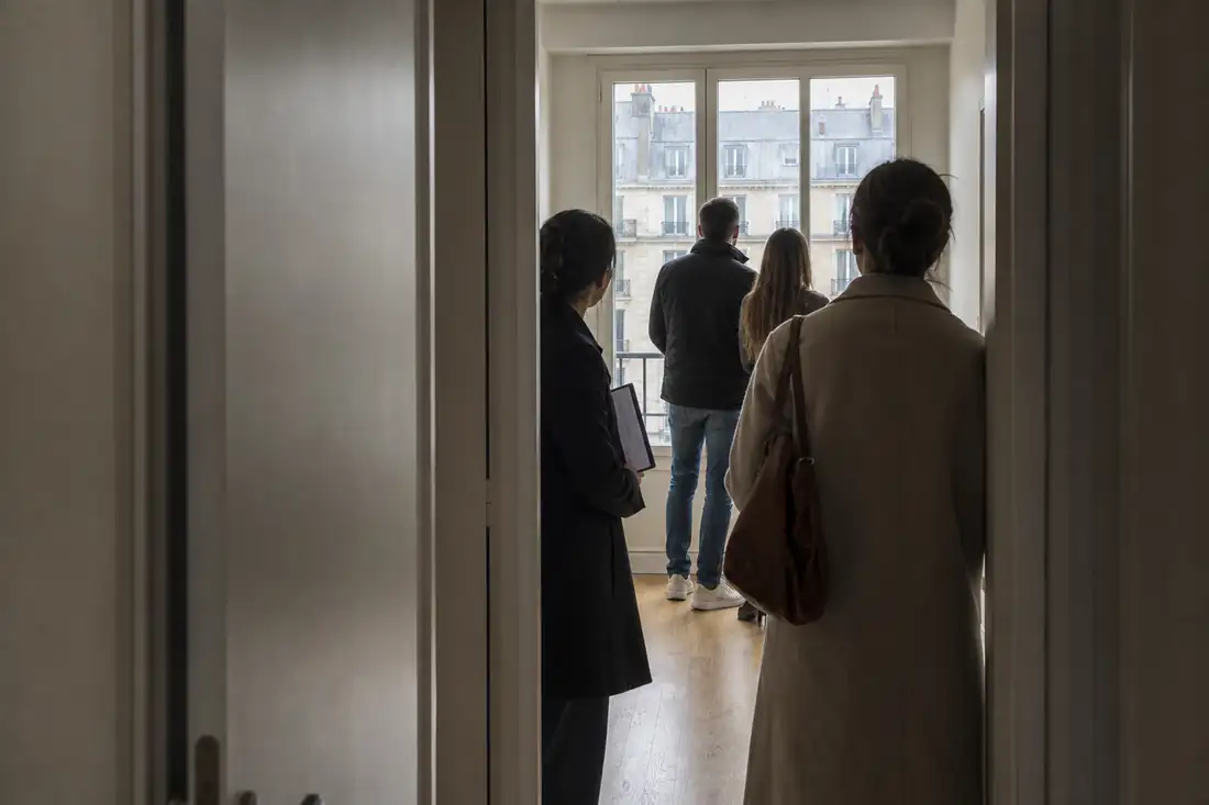 Visite d’un petit appartement en ville par plusieurs candidats locataires, près d’une grande fenêtre.