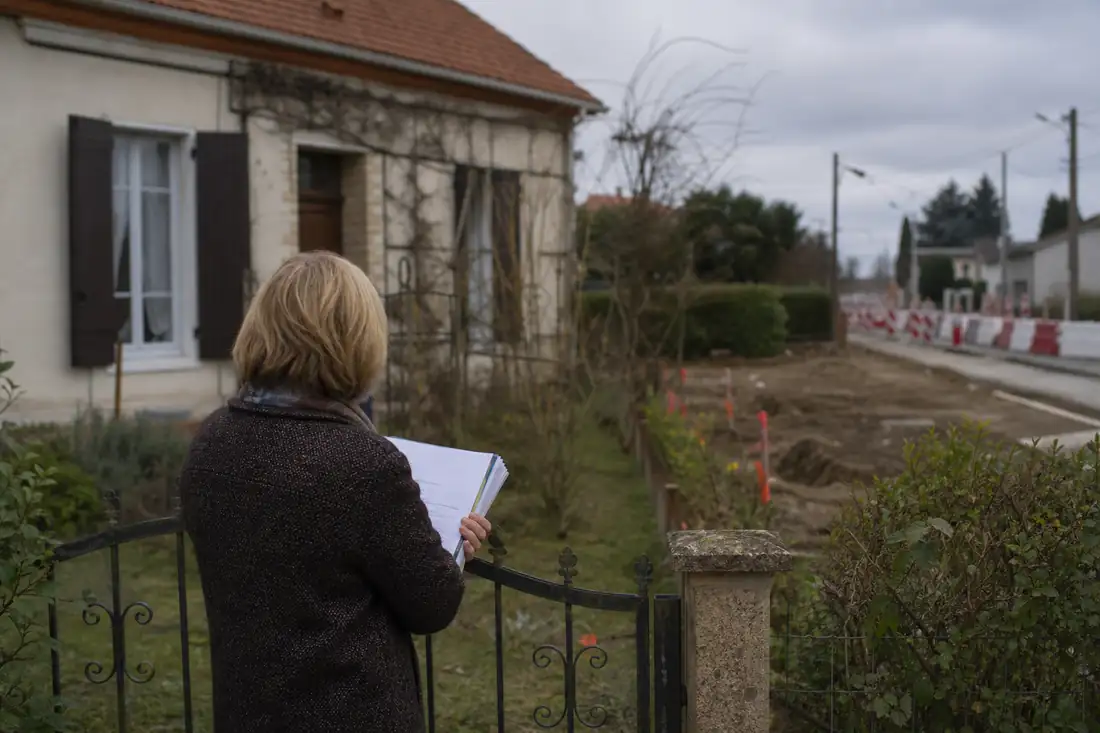 Pavillon individuel près d’un projet de voirie, avec un dossier foncier tenu devant le portail.
