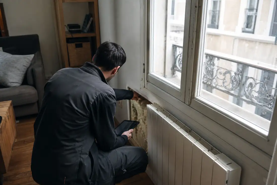 Diagnostiqueur examinant un radiateur électrique et une fenêtre dans un appartement avant mise à jour du DPE.