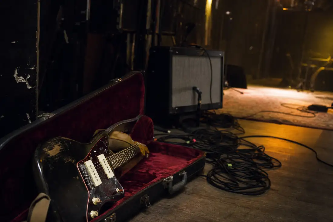Guitare électrique posée près d’un ampli et de câbles dans les coulisses d’une salle de concert vide.