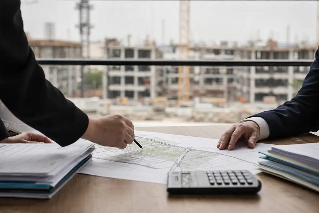 Dossier foncier et plans de construction examinés dans une base vie face à un chantier urbain.