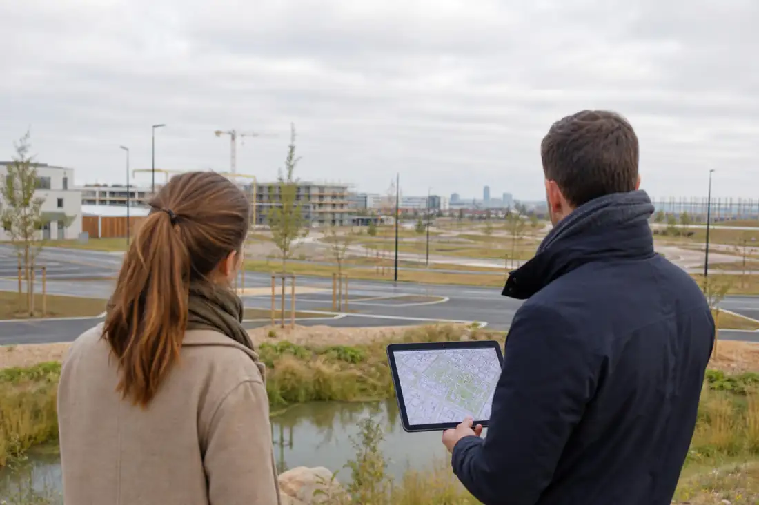 Aménagement d’un quartier en développement avec voirie neuve, parcelles préparées et immeubles au loin.