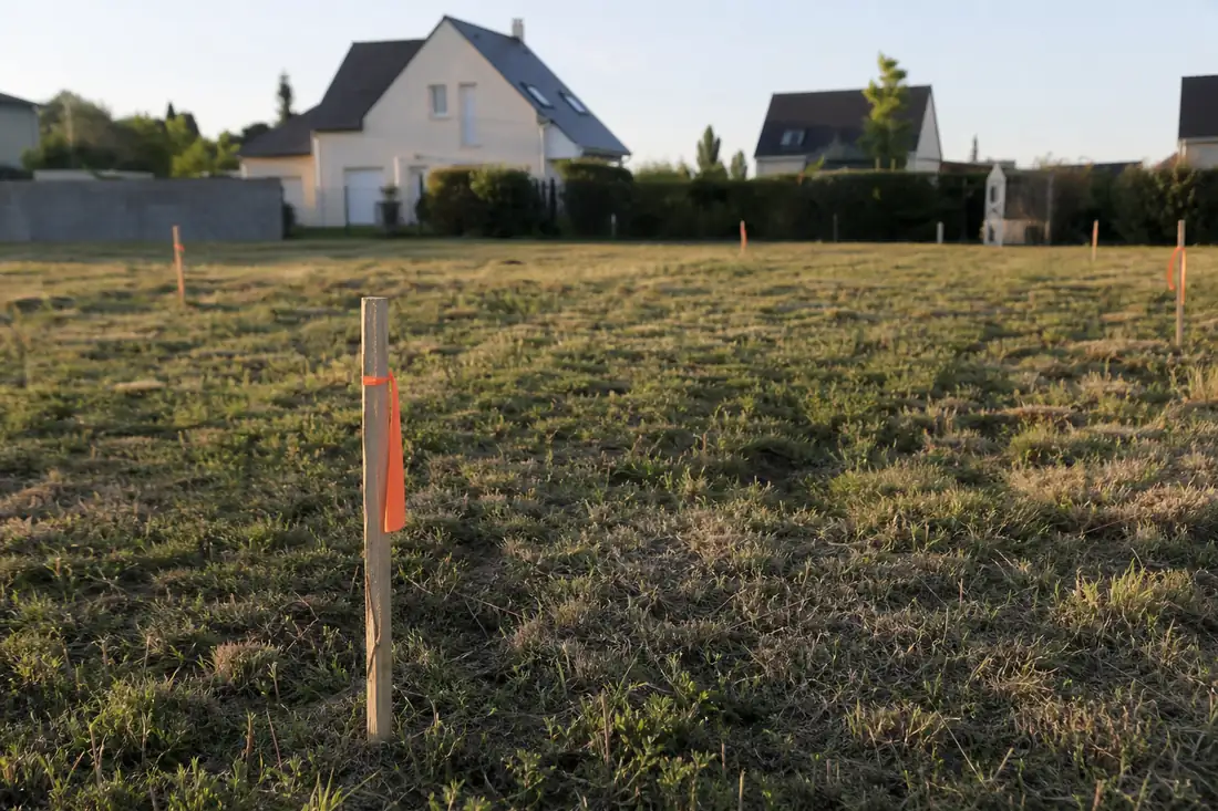 Terrain à bâtir en lotissement délimité par des piquets de géomètre, maisons individuelles voisines en arrière-plan