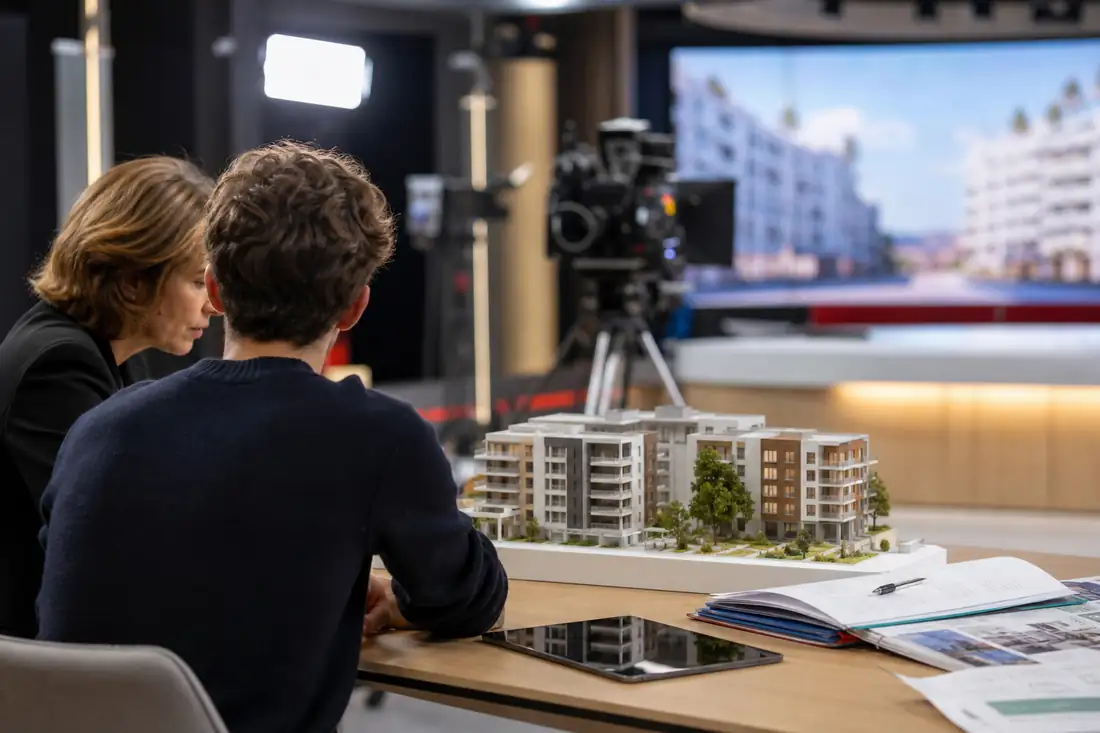 Plateau télévisé consacré au logement avec maquette d’immeuble et dossiers immobiliers posés sur une table.